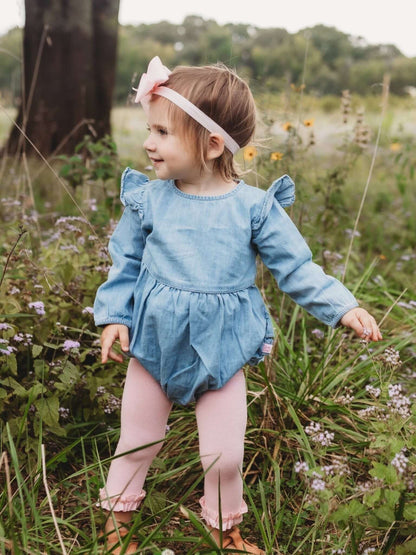 Baby girl wearing a light wash long sleeve flutter bubble romper by RuffleButts + RuggedButts, showing the adorable design and soft fabric.