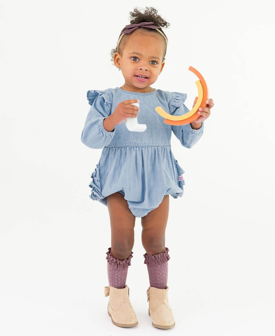 Baby girl wearing a light wash long sleeve flutter bubble romper by RuffleButts + RuggedButts, showing the cute, comfortable design.