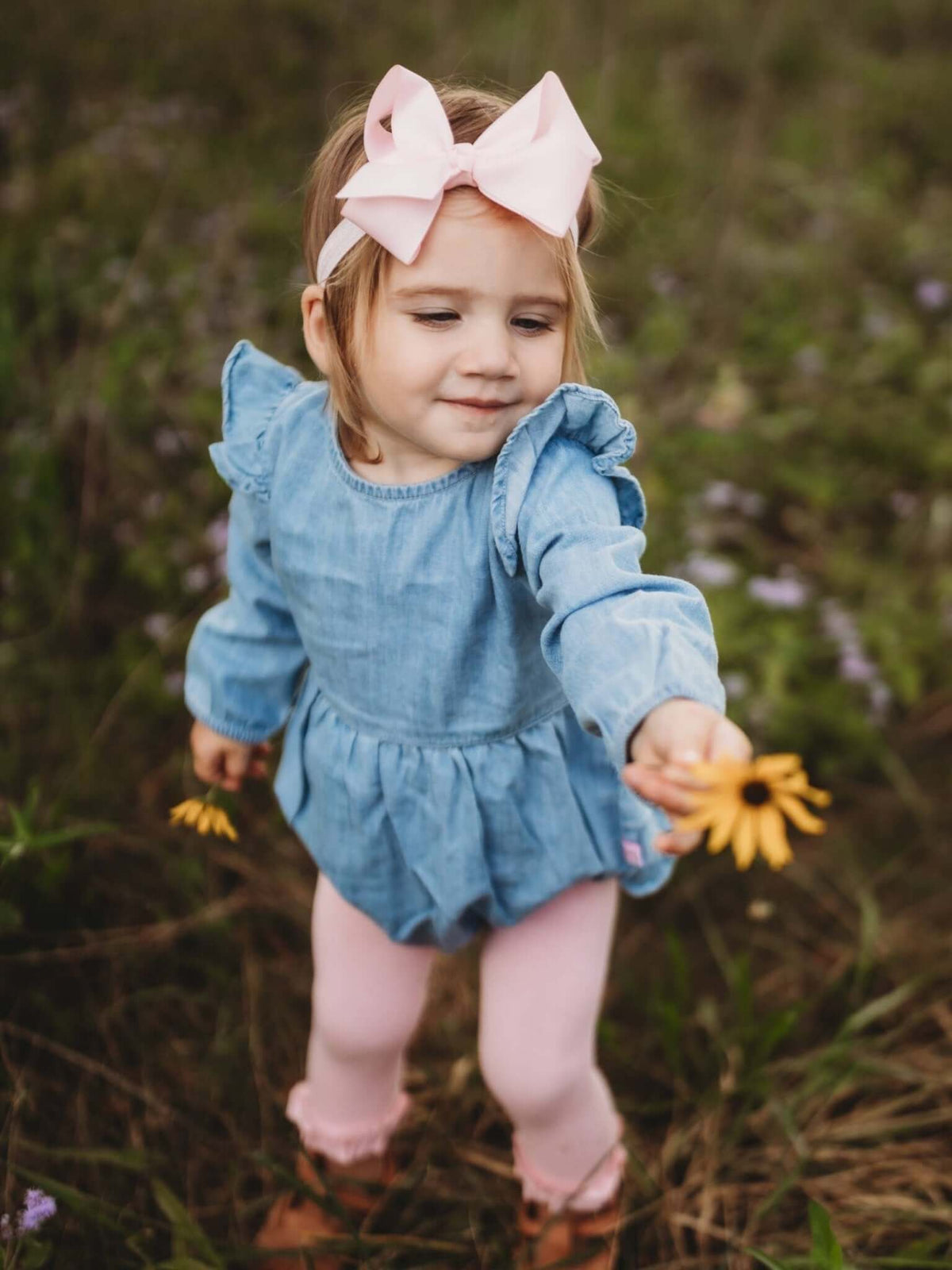 Baby girl wearing light wash long sleeve flutter bubble romper from RuffleButts + RuggedButts, showcasing adorable design and comfortable fit.
