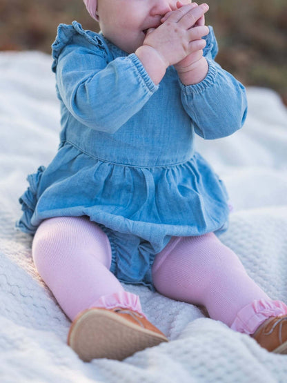 Baby girl wearing light wash long sleeve flutter bubble romper by RuffleButts + RuggedButts, showing soft fabric and cute flutter details.