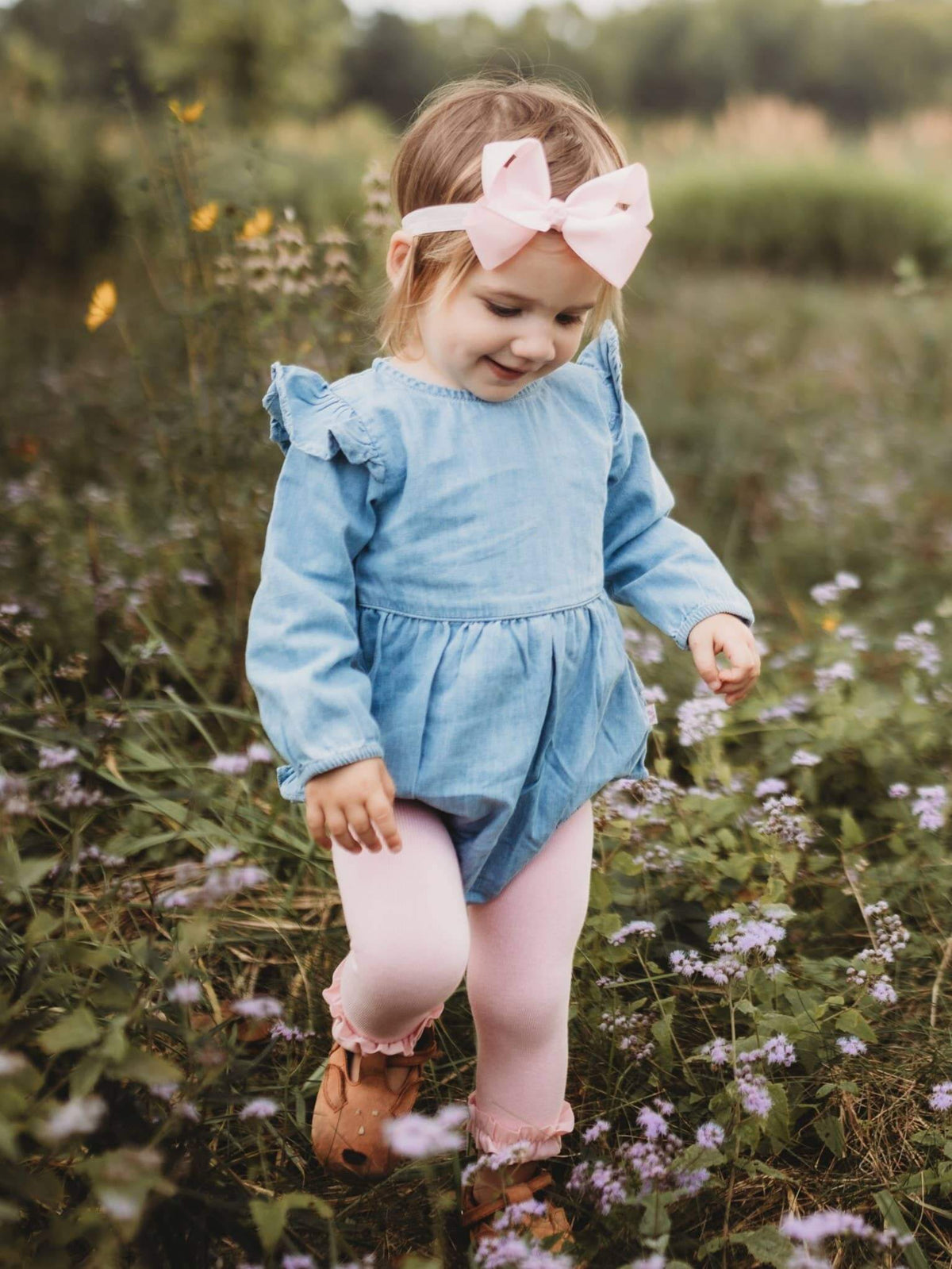 Baby girl wearing a light wash long sleeve flutter bubble romper from RuffleButts + RuggedButts, showing the cute, airy design and soft fabric.