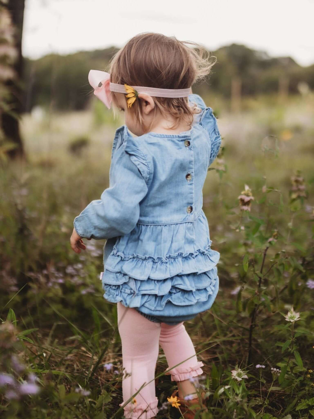 Baby girl in light wash long sleeve flutter bubble romper by RuffleButts + RuggedButts, showing the cute design and soft fabric.